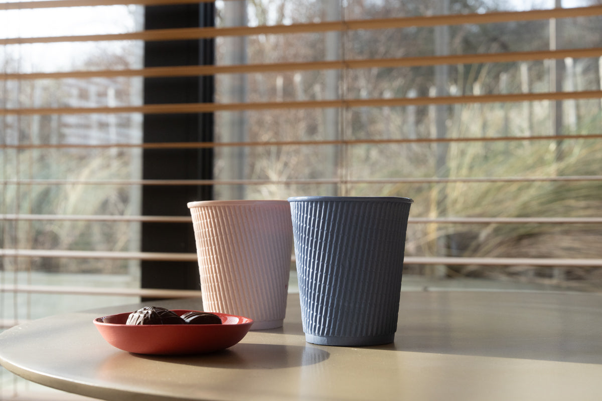 Zwei handgemache Barista Latte Kaffebecher und ein Tellerchen aus Porzellan stehen auf einem Tisch - im Hintergrund sind Dühnen und Gräser.